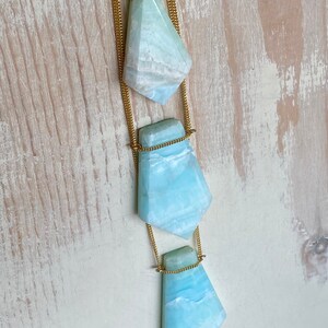Pu&ograve; includere: Una collana a strati con tre pendenti in pietra preziosa azzurra, ciascuno con un design sfaccettato. I pendenti sono sospesi a sottili catene dorate. Le pietre preziose hanno un effetto marmorizzato con tonalit&agrave; bianche e blu. La collana &egrave; posta su uno sfondo chiaro e strutturato.