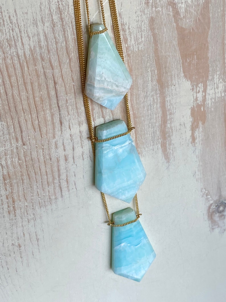 Pu&ograve; includere: Una collana a strati con tre pendenti in pietra preziosa azzurra, ciascuno con un design sfaccettato. I pendenti sono sospesi a sottili catene dorate. Le pietre preziose hanno un effetto marmorizzato con tonalit&agrave; bianche e blu. La collana &egrave; posta su uno sfondo chiaro e strutturato.