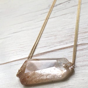May include: A gold chain necklace with a large, clear, faceted crystal pendant. The crystal has brown inclusions that resemble hair.