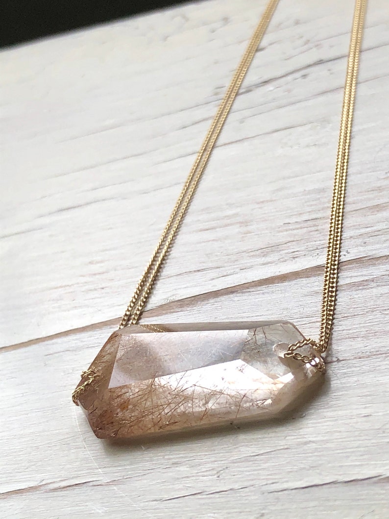 May include: A gold chain necklace with a large, clear, faceted crystal pendant. The crystal has brown inclusions that resemble hair.