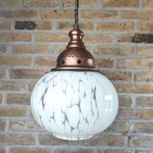 May include: A vintage pendant light fixture with a white globe shade featuring a gray, speckled pattern. The fixture has a copper-colored top and a chain for hanging. The light is suspended against a brick wall.
