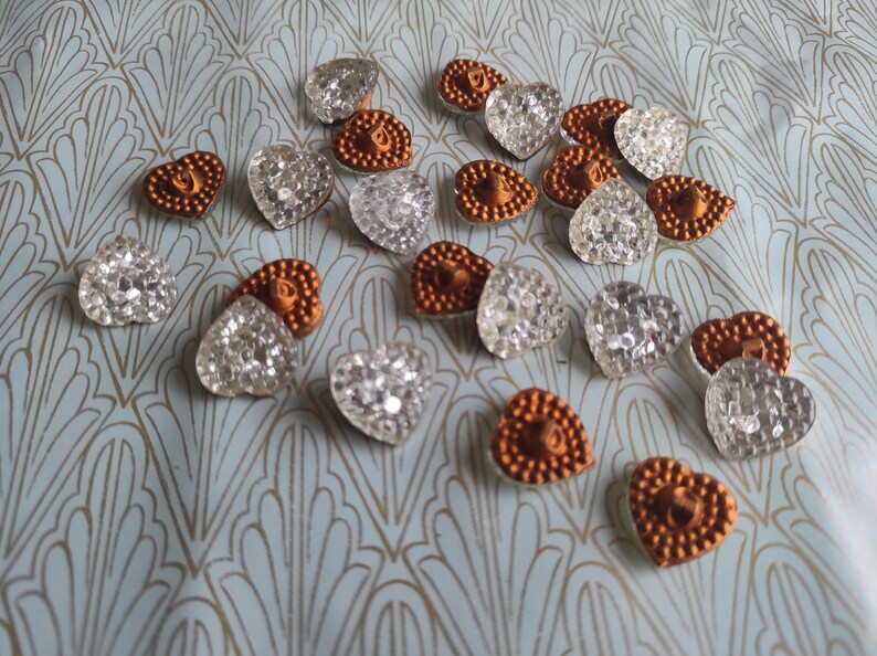 May include: A collection of heart-shaped buttons in two colors: clear and copper. The buttons are arranged on a blue and gold patterned surface.