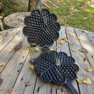 Può includere: Una macchina per waffle in ghisa nera con un design a forma di cuore, aperta e appoggiata su una superficie di legno. La macchina per waffle è aperta, rivelando l'interno a motivi. Il manico è lungo e nero. Lo sfondo include un tavolo di legno e fogliame.