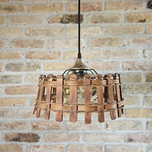 May include: A rustic pendant light with a woven wooden shade. The shade is made of vertical wooden slats held together by horizontal woven strips. The light fixture has a brass-colored top and a black cord, hanging against a brick wall.