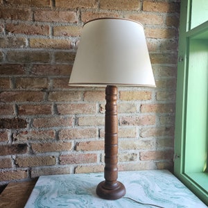 Könnte beinhalten: Eine Tischlampe mit einem gedrechselten Holzfuß in Dunkelbraun und einem zylindrischen, cremeweißen Stoffschirm. Die Lampe hat ein schlichtes, vertikales Design mit einem runden Sockel und einer glatten, polierten Oberfläche am Holzständer.
