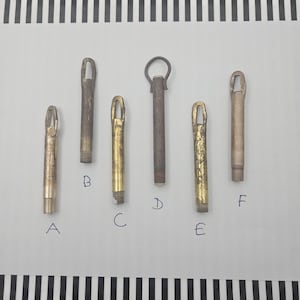 Può includere: Un assortimento di sei tiranti in metallo vintage, ognuno con un design unico. I tiranti variano di colore, dall'oro ottone al bronzo invecchiato. Ogni tirante ha una base filettata e un anello o anello nella parte superiore.