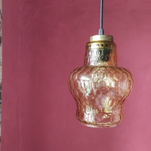 Puede incluir: Lámpara colgante de estilo vintage con una pantalla de cristal ámbar texturizado. La pantalla tiene forma bulbosa y una parte superior de metal color latón. La luz cuelga de un cable negro contra una pared rosa.
