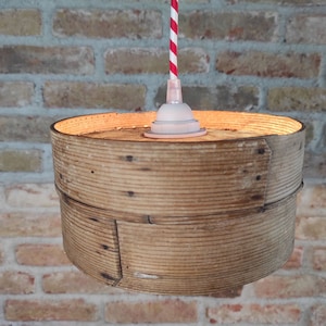 Puede incluir: Lámpara colgante cilíndrica rústica de madera clara. La lámpara tiene un interior cálido y brillante y está suspendida por un cable a rayas rojas y blancas. El fondo es una pared de ladrillos.