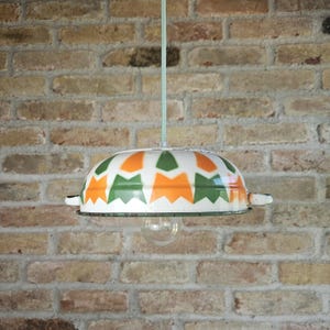 May include: A vintage-style pendant light with a white, orange, and green patterned shade. The shade is bowl-shaped with a decorative edge and small handles. The light hangs from a light green cord against a brick wall.