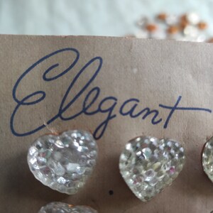 May include: A card with the word "Elegant" handwritten in black ink. The card has several clear glass heart-shaped buttons attached to it.