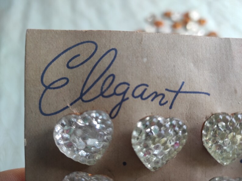 May include: A card with the word "Elegant" handwritten in black ink. The card has several clear glass heart-shaped buttons attached to it.