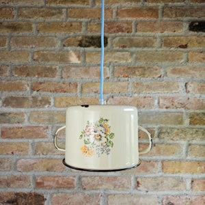 May include: Cream-coloured, vintage-style pendant light. The light is made from a repurposed pot with a floral design. The pot has two handles and a black rim. A light blue cord suspends the light against a brick wall.