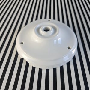 May include: A white ceramic ceiling canopy with a central hole and three smaller holes around the edge. The canopy has a rounded shape and a glossy finish. The background is a black and white striped pattern.