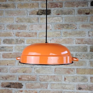 May include: Orange pendant light fixture with a black rim and a black cord. The light has a rounded shape with small handles on each side. The background is a brick wall.