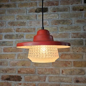 May include: A red pendant light fixture with a circular shade and a patterned glass diffuser. The light hangs from a black cord against a brick wall. The fixture has a vintage or retro aesthetic.