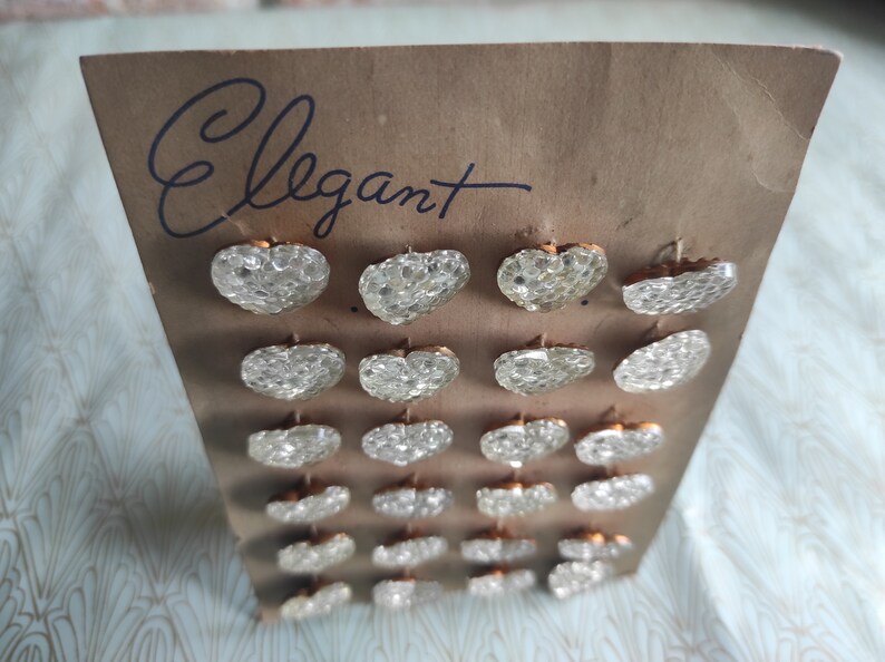 May include: A card with the word "Elegant" written on it. The card has 20 clear heart-shaped buttons with gold-colored backs.