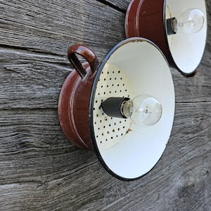 Peut inclure: Deux appliques murales de style vintage fabriquées à partir de passoires de cuisine recyclées. Les appliques sont marron avec des intérieurs blancs et des bordures noires. Chaque applique a une poignée et une ampoule. Le fond est une surface en bois vieilli.