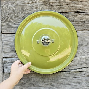 Peut inclure: Un luminaire rond en métal vert olive avec un bord noir et une ampoule transparente au centre. Le luminaire est monté sur une surface en bois vieilli. Une main tient le bord du luminaire.