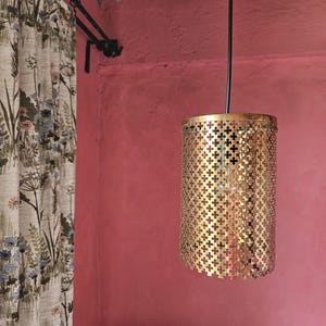 May include: A gold pendant light fixture with a geometric pattern hangs from a black cord against a pink wall. The light fixture is made of metal and has a cylindrical shape.