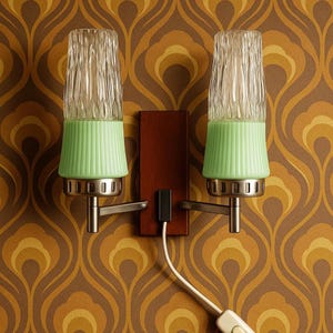 May include: A pair of vintage wall sconces with a retro design. Each sconce features a clear, textured glass shade atop a light green base and a brushed metal arm. The sconces are mounted on a dark wood backing, with a white cord and switch.