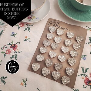 May include: A collection of clear glass heart-shaped buttons arranged on a brown card with the word "Elegant" written on it. The buttons are arranged in rows and are all the same size and shape.
