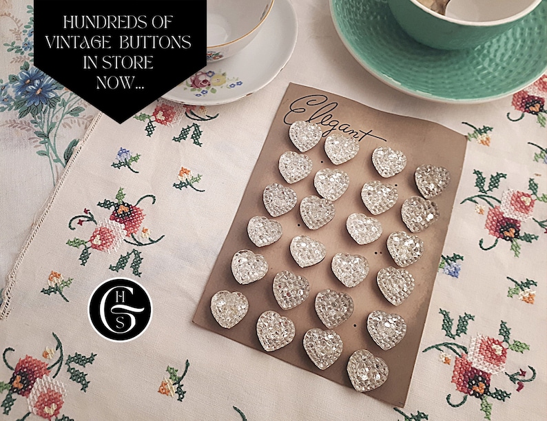 May include: A collection of clear glass heart-shaped buttons arranged on a brown card with the word "Elegant" written on it. The buttons are arranged in rows and are all the same size and shape.