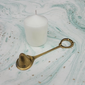 Puede incluir: Una vela blanca cilíndrica junto a un apagavelas de latón. El apagavelas tiene un cuenco en forma de cuchara y un mango largo con un anillo en el extremo. La vela y el apagavelas están sobre una superficie marmolada.