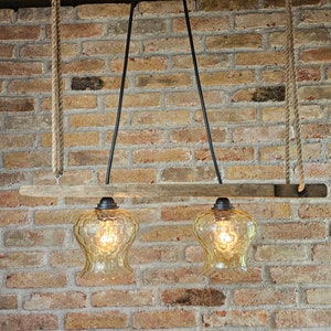 May include: A rustic, two-light pendant lamp with amber-colored glass shades. The lamp is suspended by rope and black metal rods from a wooden beam. The background is a brick wall, creating a warm, inviting ambiance. The light fixtures are illuminated.
