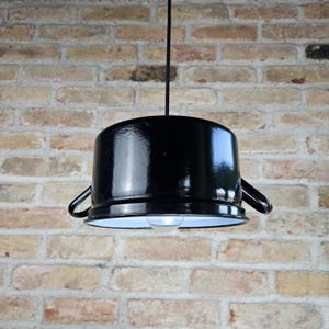 Puede incluir: Luminaria colgante de olla de esmalte negro con interior blanco y una sola bombilla. La luminaria está colgada de un cable negro contra una pared de ladrillo.