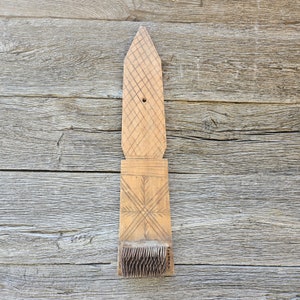 May include: A vintage wooden wool comb with a long, pointed handle and a rectangular base with metal teeth. The handle features carved diamond and crosshatch patterns. The comb is made of light-colored wood and is set against a weathered wood background.