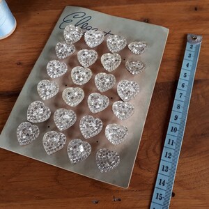 May include: A sheet of clear heart-shaped buttons with a sparkly finish. The buttons are arranged in rows on a light gray card with the word "Elegant" written in black ink.