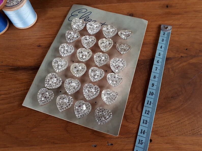 May include: A sheet of clear heart-shaped buttons with a sparkly finish. The buttons are arranged in rows on a light gray card with the word "Elegant" written in black ink.