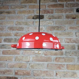 Op de afbeelding: Rode hanglamp met witte stippen en een zwart snoer dat aan een bakstenen muur hangt.