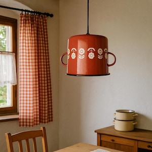Può includere: Una lampada a sospensione rossa con un motivo floreale bianco e due maniglie laterali. La luce è appesa a un cavo nero. L'apparecchio si trova in una stanza con una finestra e una tenda a scacchi rossi e bianchi.