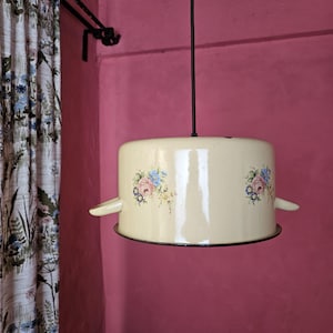 Puede incluir: Lámpara colgante de estilo vintage color crema. La lámpara tiene forma de olla con dos asas laterales y calcomanías florales en rosa, azul y amarillo. La lámpara cuelga de un cable negro contra una pared rosa.