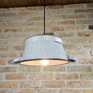 May include: A white metal pendant light fixture with a black cord and a light bulb. The fixture is shaped like a large pot with two handles.