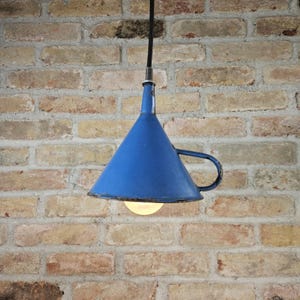 Puede incluir: Una lámpara colgante de metal azul en forma de embudo cuelga de un cable negro contra una pared de ladrillo.