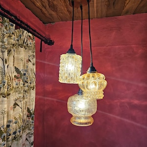 May include: Three vintage pendant lights with textured, amber-coloured glass shades hang from a wooden ceiling fixture. The shades have unique patterns and shapes, casting a warm glow against a red wall.