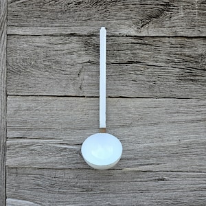 May include: A white enamel ladle with a long, straight handle and a deep, rounded bowl. The ladle features a smooth, glossy white finish throughout, with a small metallic band connecting the handle to the bowl, presenting a clean, functional design.