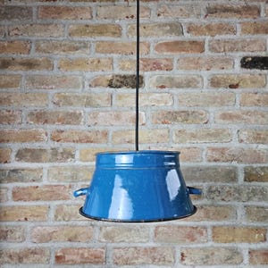 May include: A vintage blue enamel pendant light fixture with a black cord, hanging against a brick wall. The light has a flared shape with small handles on each side. The blue enamel has a glossy finish.