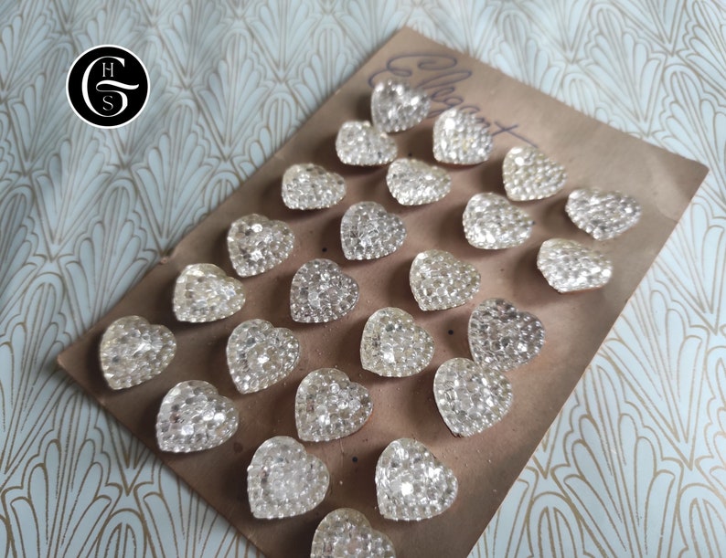 May include: A collection of 20 clear glass heart-shaped embellishments on a brown surface. The embellishments are arranged in a pattern and are all the same size.