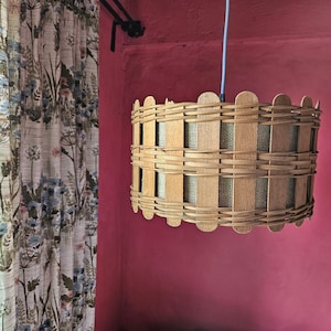 May include: A woven pendant light fixture with a cylindrical shape. The light is made of light brown wood strips woven together, with a beige fabric lining visible through the openings. It hangs from a blue cord against a pink wall.