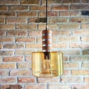 Può includere: Un lampadario a sospensione con un paralume cilindrico in vetro ambrato e una parte superiore in legno e metallo. La lampadina è visibile all'interno del paralume. L'apparecchio è appeso a un cavo nero contro un muro di mattoni.