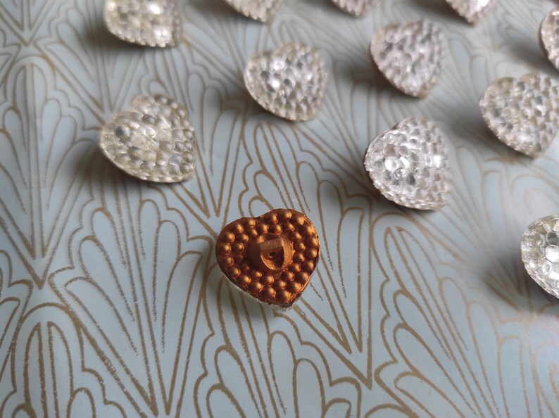 May include: A collection of heart-shaped buttons with a gold-colored button in the center. The buttons are made of a shiny, textured material and have a raised design.