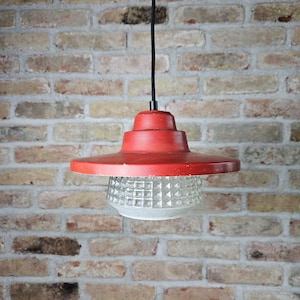 May include: A red, vintage pendant light with a wide, flat shade and a textured glass globe. The light hangs from a black cord against a brick wall. The light's dimensions are approximately 30 cm in diameter.