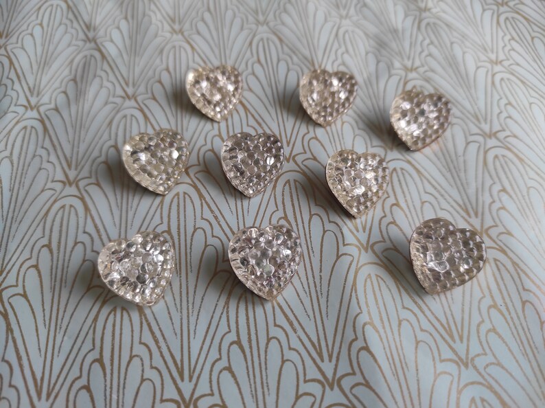 May include: Nine silver heart-shaped buttons with a textured, sparkly finish. The buttons are arranged in a semi-circle on a light blue background with a gold pattern.