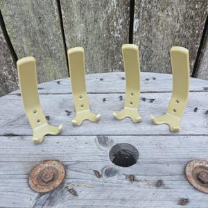 Può includere: Quattro ganci da parete color beige chiaro disposti in fila. Ogni gancio presenta una piastra posteriore verticale curva con due fori di montaggio circolari e una base a due punte per appendere oggetti. Finitura liscia e tinta unita.