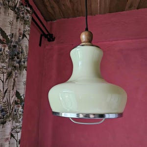 May include: A pale yellow glass pendant light with a wooden top and a chrome trim. The light is hanging from a black cord against a red wall.