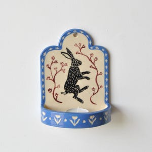 May include: A wall-mounted ceramic candle holder with a cream-colored back panel featuring a black hare illustration and red floral vines. The frame is blue with white diamond accents, and the base features a white floral pattern on blue.