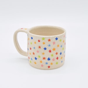 Könnte beinhalten: Cremefarbene Keramiktasse mit Henkel, verziert mit einem Muster aus bunten Sternen. Die Sterne sind in den Farben Rot, Orange, Gelb, Grün, Blau und Rosa gehalten. Die Tasse ist für heiße oder kalte Getränke geeignet.